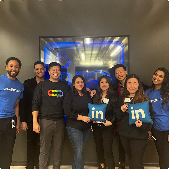 Group of eight diverse individuals at a LinkedIn event, smiling and posing for a photo indoors, some holding blue LinkedIn pillows, with a large digital display in the background.