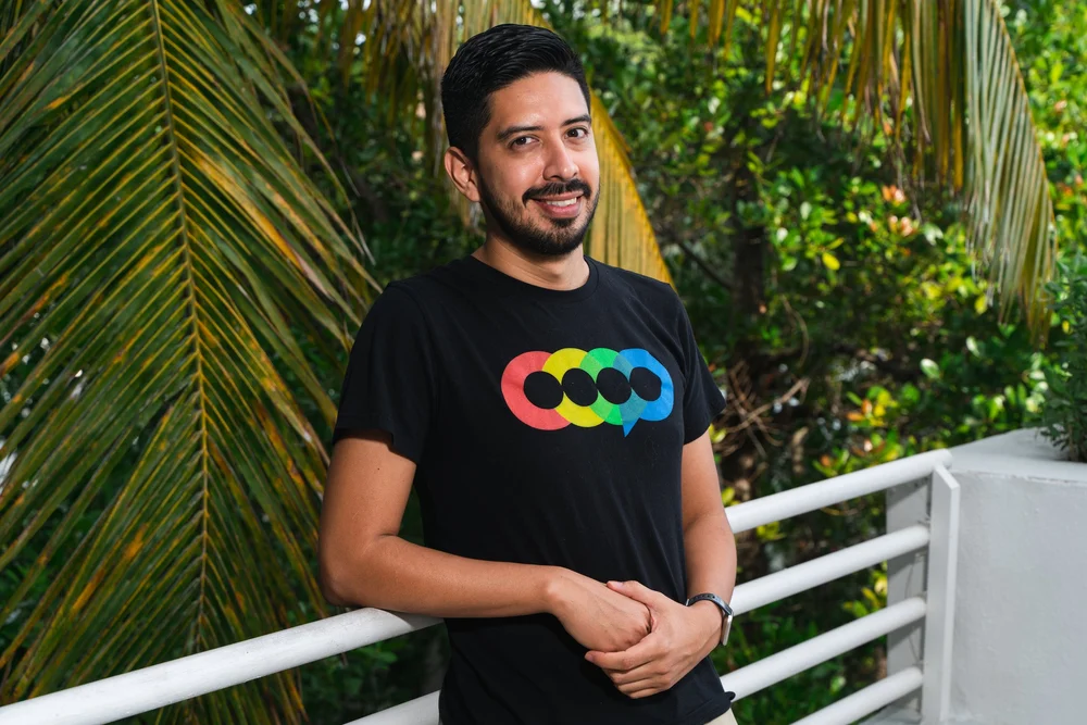 Smiling man standing outdoors, leaning on a white railing, surrounded by green tropical plants.