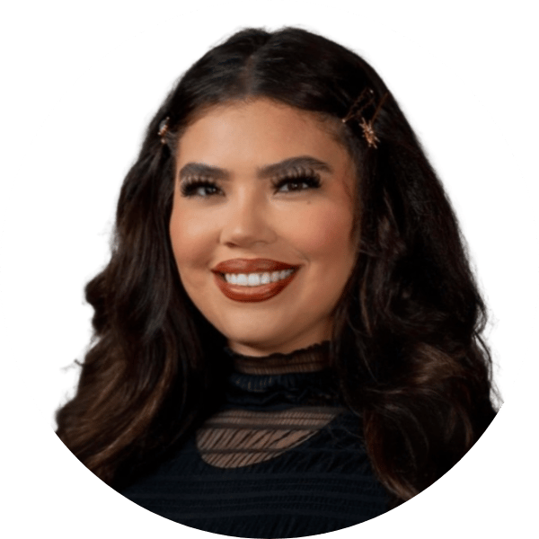 Smiling woman with dark wavy hair, hair clips, and makeup, wearing a black top with a sheer neckline against a white background.