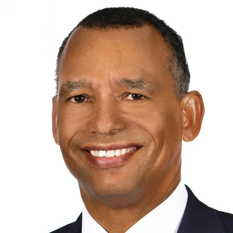 Close-up snapshot of a smiling man in a dark suit and white shirt against a white background.