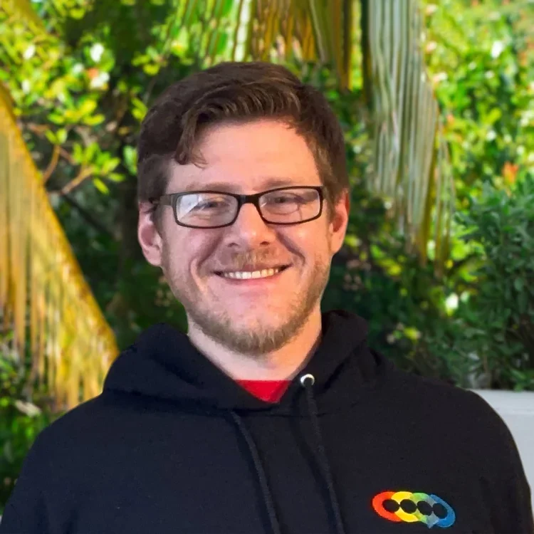 A smiling man with glasses and a beard, wearing a black hoodie with a colorful logo, standing outdoors with green foliage in the background.