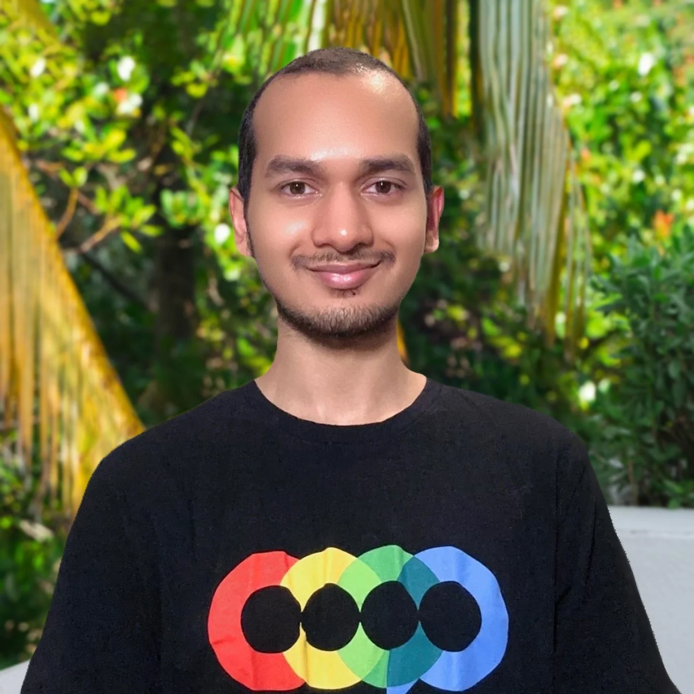 A man with short dark hair and a goatee smiling at the camera against a background of lush green plants and trees, wearing a black T-shirt with a colorful rainbow heart design.