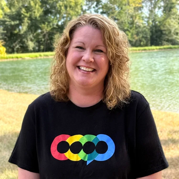 A woman with curly blonde hair smiling outdoors near a lake, wearing a black t-shirt with a colorful overlapping circle design.