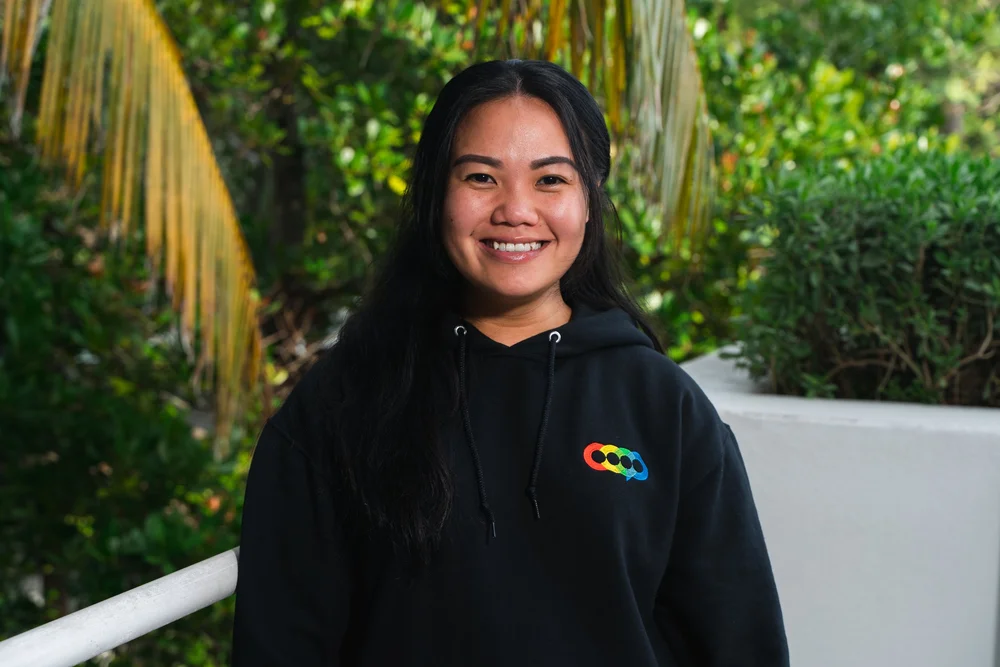 A smiling woman with long black hair wearing a black hoodie with a rainbow-colored logo, standing outdoors with green plants and palm trees behind her.