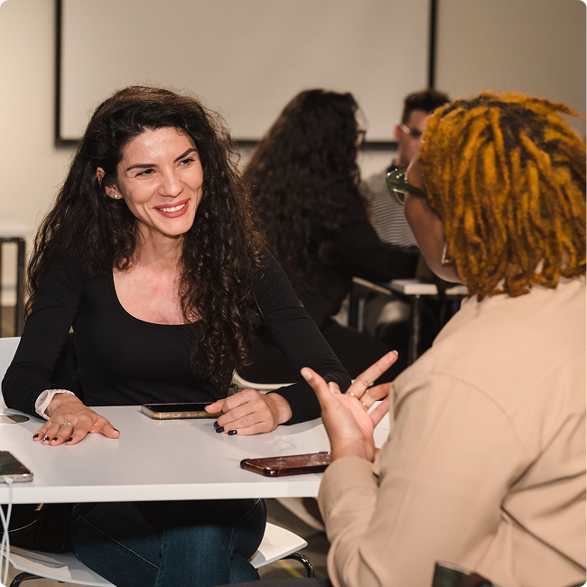 Two women engaging in conversation at a table, one with long dark curly hair and the other with short curly blond hair and glasses, in a casual indoor setting.