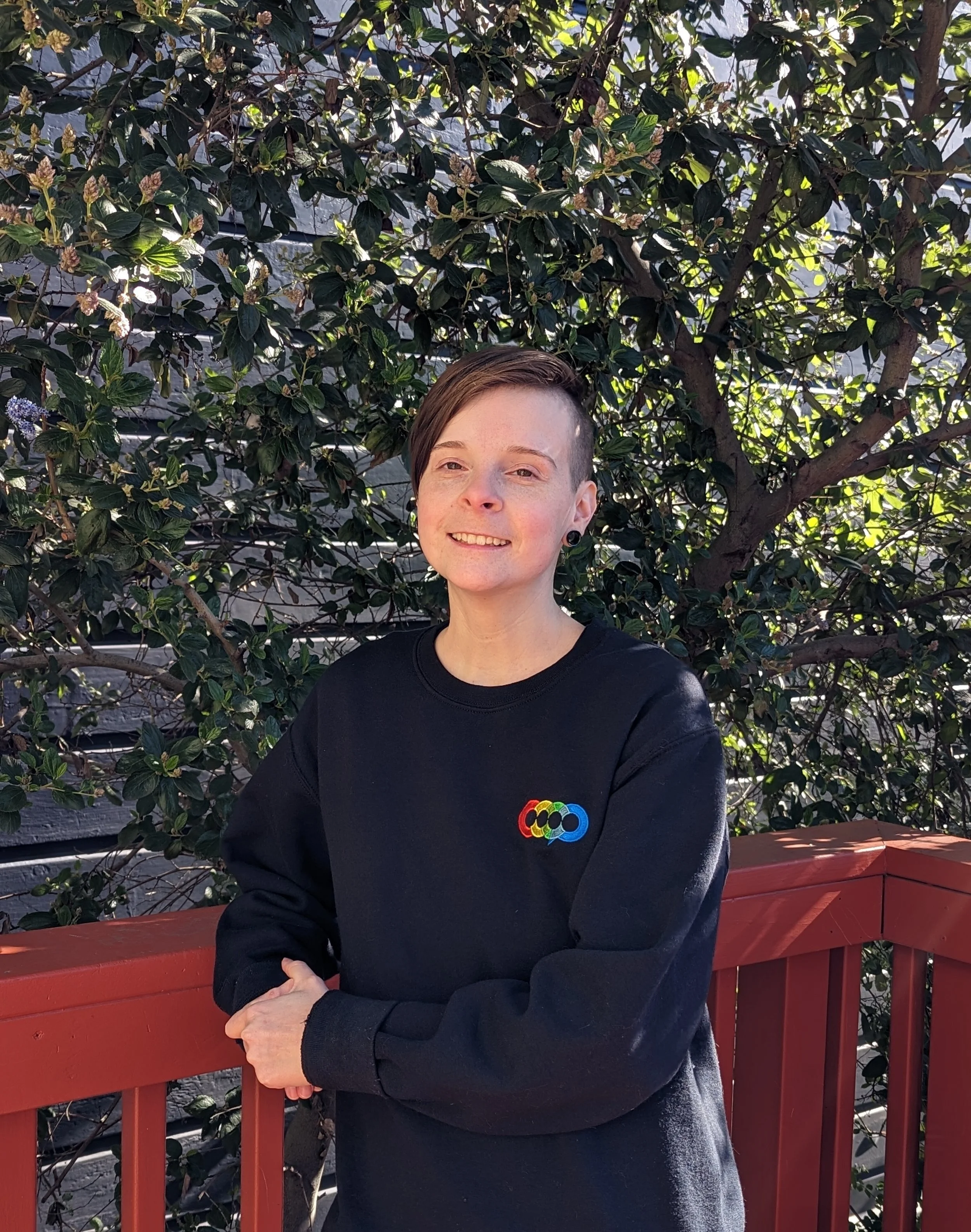 A person with short brown hair and facial piercings smiling outdoors, standing in front of a green leafy tree and a red wooden railing.