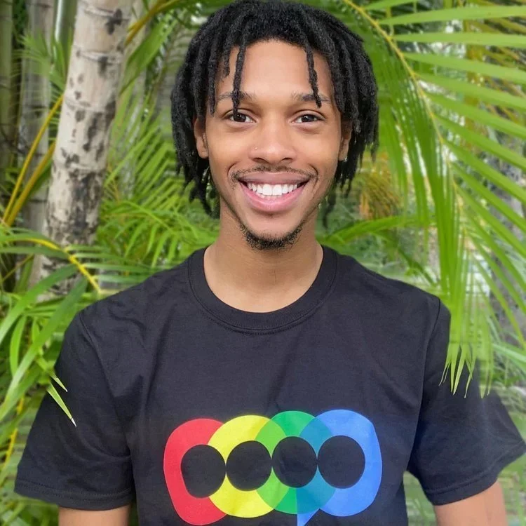 A young man with curly dreadlocks smiling, wearing a black t-shirt with a colorful logo, standing outdoors among green plants.