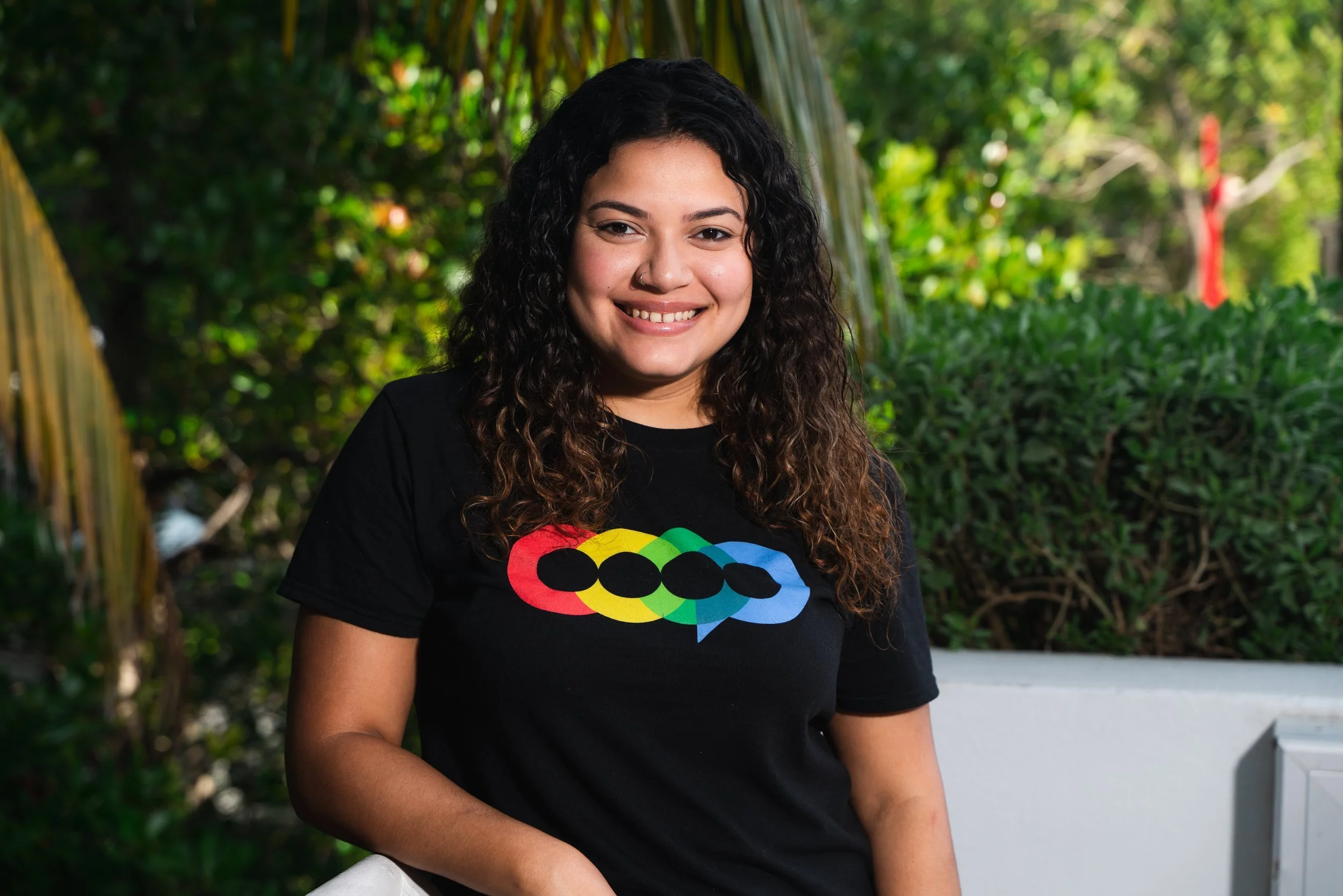 A young woman with curly dark hair and a nose ring smiling outdoors. She is wearing a black T-shirt with an LGBTQ+ pride flag speech bubble graphic. Background includes trees and greenery.