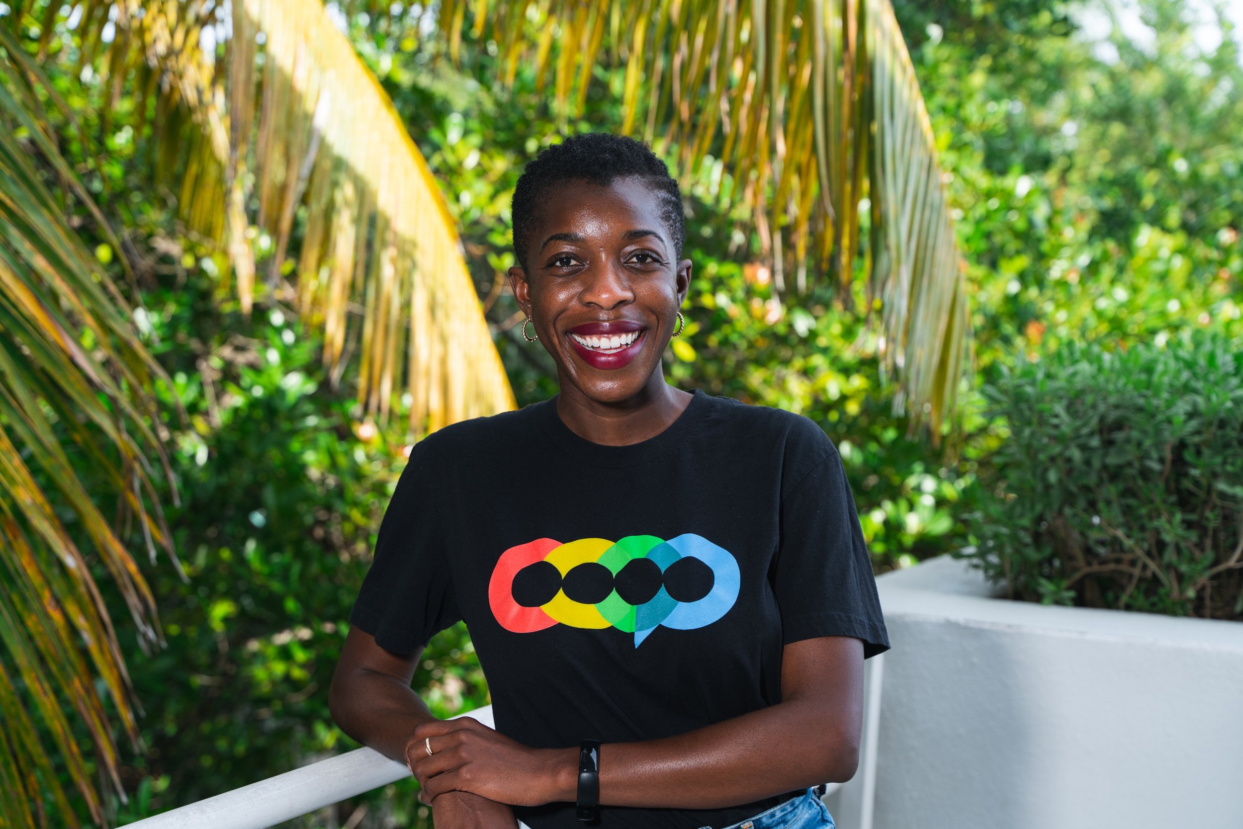 Smiling woman with short curly hair and earrings, wearing a black t-shirt with rainbow-colored speech bubbles, standing outdoors near palm trees and green foliage.