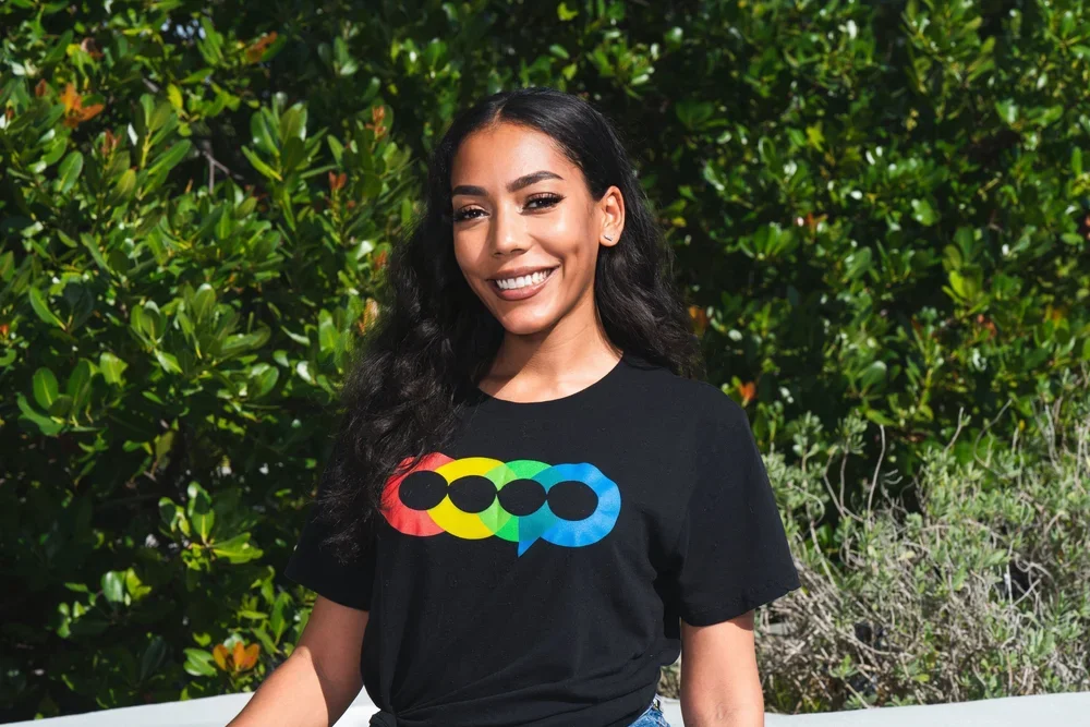 A young woman with long dark hair smiling outdoors, wearing a black T-shirt with a colorful print design, standing next to lush green shrubbery.