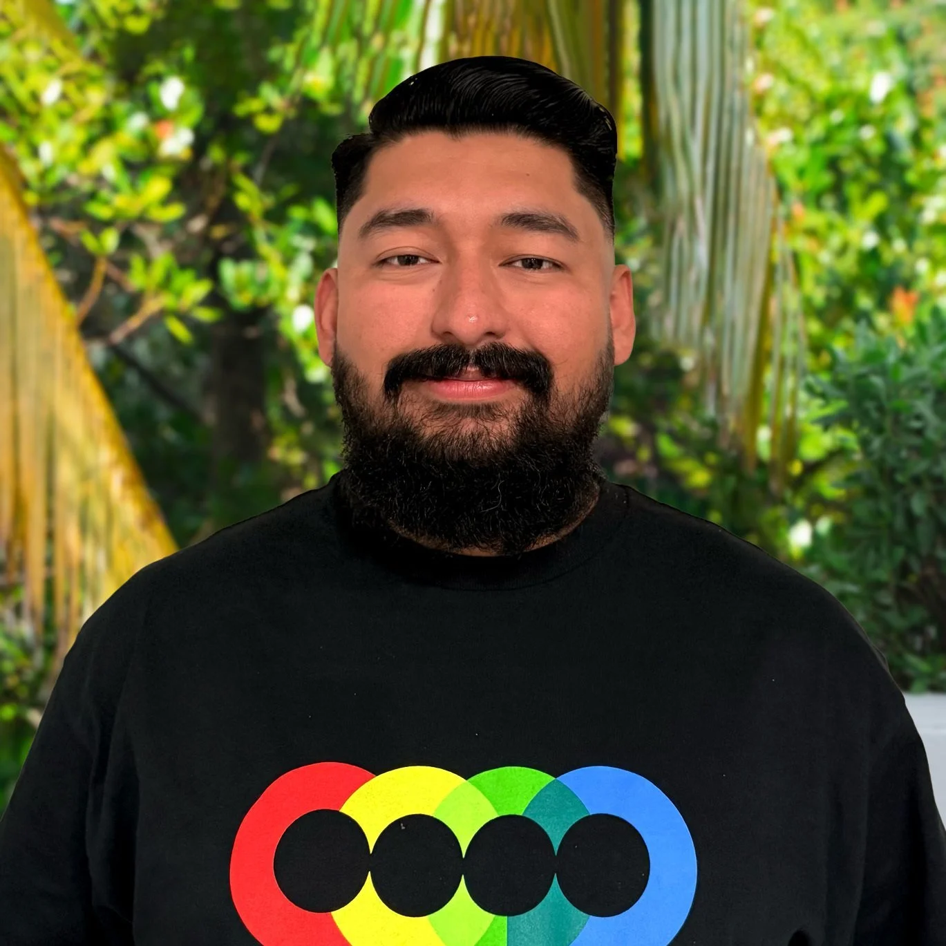 A man with a dark beard and dark hair, wearing a black shirt with a colorful rainbow logo, stands in front of a green leafy background.