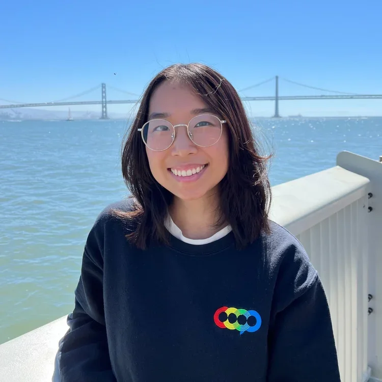 A smiling young woman with glasses and shoulder-length dark hair, standing near water with a bridge in the background on a clear, sunny day.