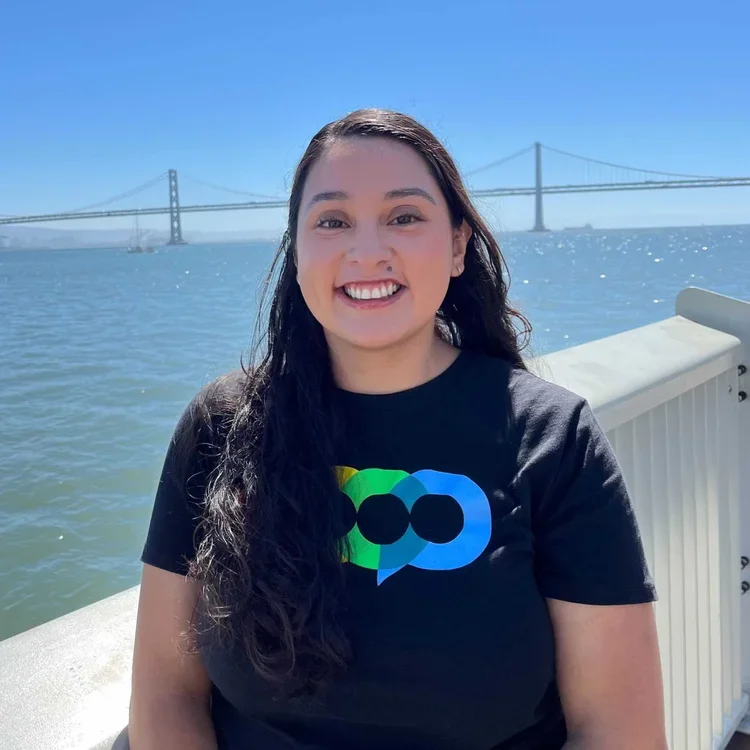 A young woman smiling outdoors near a body of water, with a bridge in the background under a clear blue sky.