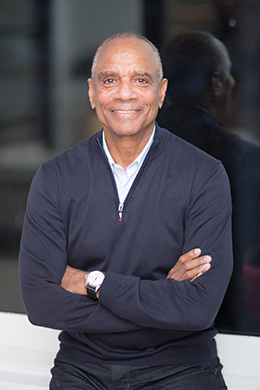 A smiling middle-aged man in a navy blue sweater with a collared shirt, standing with arms crossed in front of a window with a reflection.