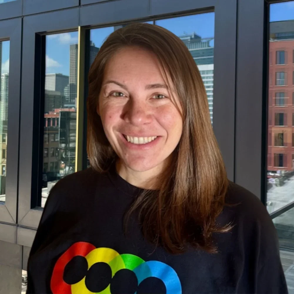 A woman with long brown hair, smiling, wearing a black shirt with a colorful rainbow-colored LGBTQ+ pride flag design, standing inside a building with large windows showing city buildings outside.