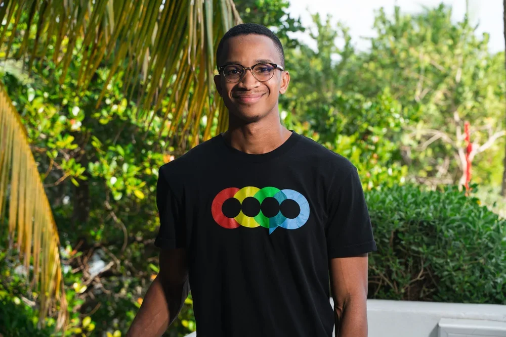 Young man wearing glasses and a black T-shirt with colorful circular and speech bubble design, standing outdoors with greenery in the background.