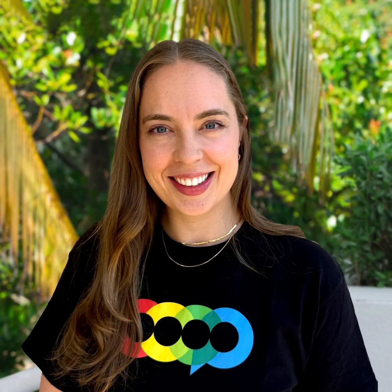 A woman with long brown hair, wearing a black shirt, smiling against a tropical background.