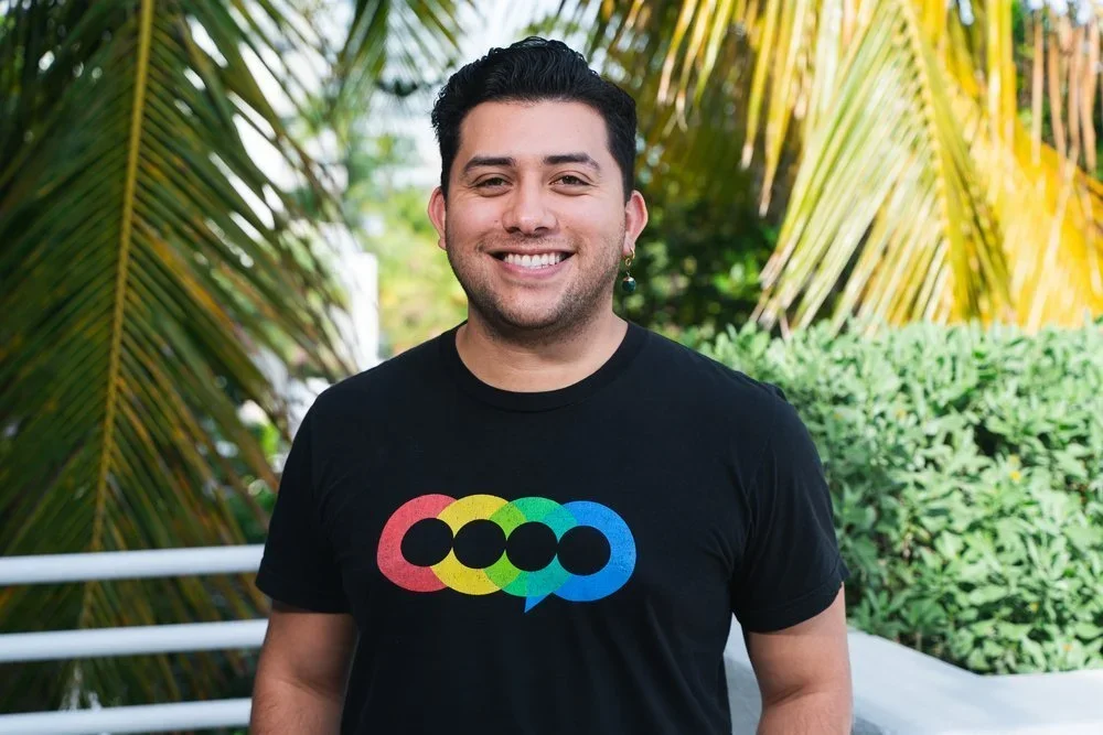 A smiling man with dark hair and earrings, wearing a black T-shirt with a colorful logo, standing outdoors with palm trees and greenery in the background.