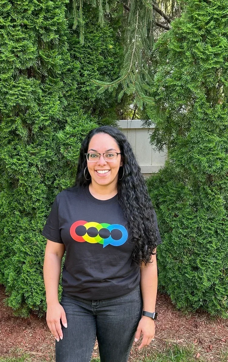 A woman with long, curly black hair and glasses smiling outdoors standing in front of green bushes.
