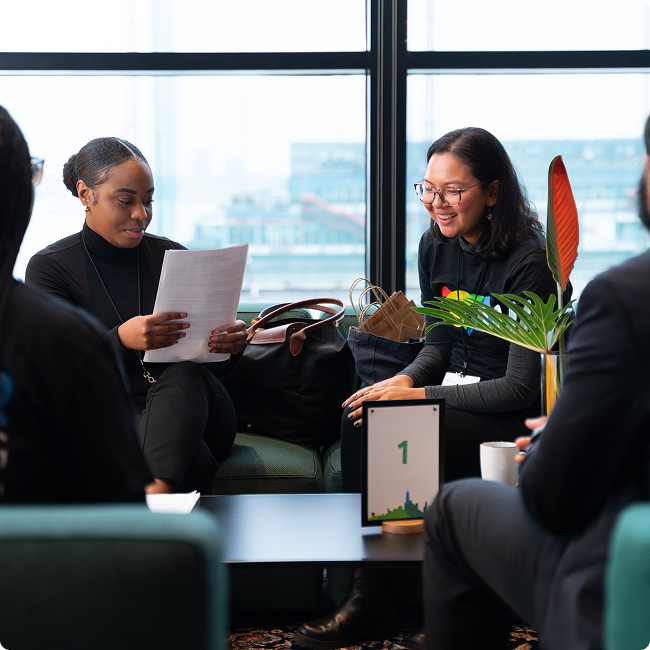 A group of diverse people having a conversation in a modern office lounge, with large windows showing a cityscape outside.