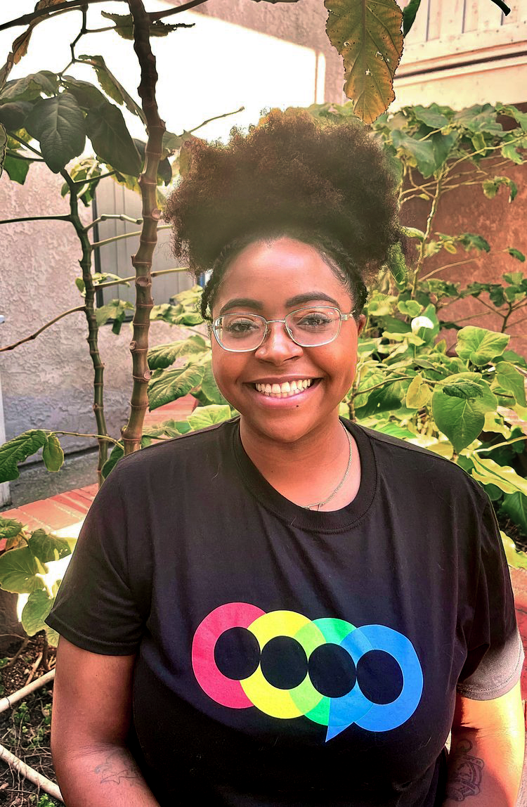 Smiling woman with glasses and a black T-shirt featuring colorful speech bubbles, standing in a garden with green plants and sunlight.