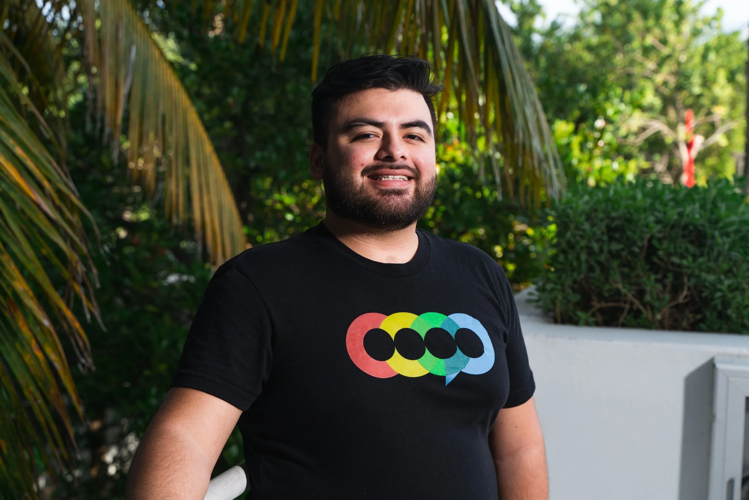 A young man with a beard and dark hair standing outdoors with greenery in the background, wearing a black T-shirt with a colorful, overlapping circles design.