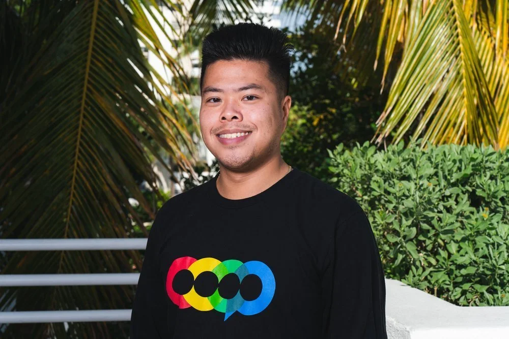 A smiling young man in a black shirt with a colorful logo stands outdoors with palm trees and green foliage in the background.