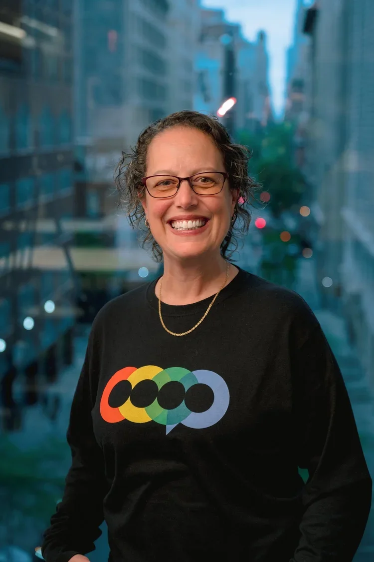 A smiling woman with curly short hair and glasses, wearing a black sweatshirt with a colorful rainbow speech bubble logo, standing in front of a cityscape with glass buildings at dusk.