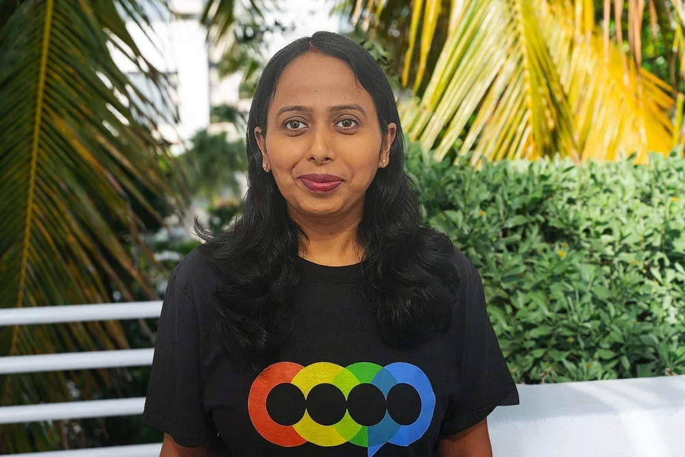 A woman with long black hair, wearing a black T-shirt with a colorful logo, standing outdoors with palm trees and green bushes in the background.