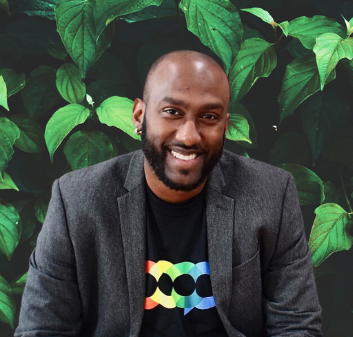 A smiling man with a beard and bald head wearing a dark blazer and a colorful graphic T-shirt, sitting in front of green leafy plants.
