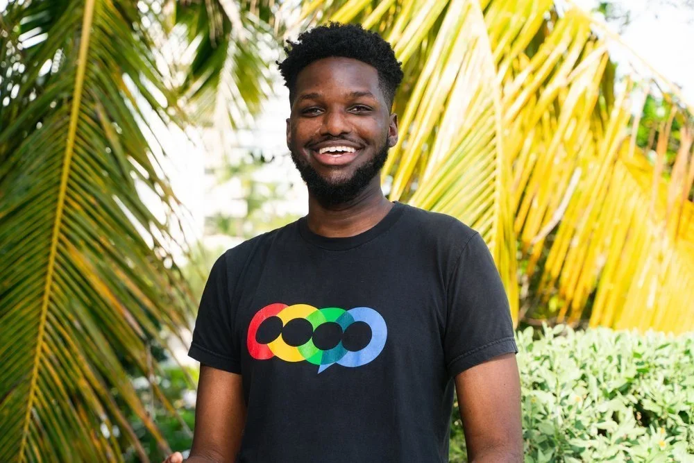 A smiling man with dark skin, black curly hair, and a beard, wearing a black T-shirt with colorful circles and speech bubble design, standing outdoors in front of lush green tropical plants and yellow palm leaves.