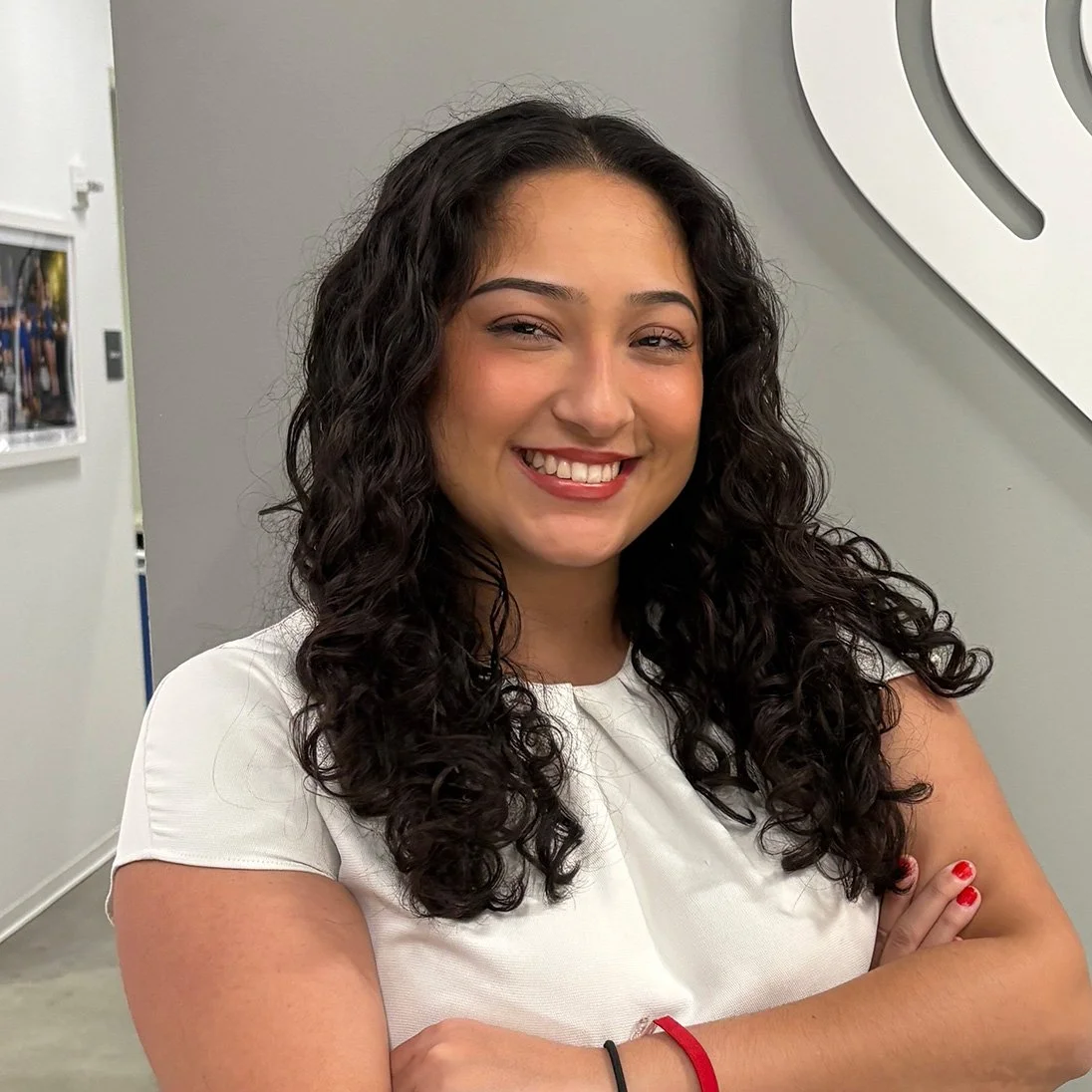 A young woman with curly dark hair smiling and crossing her arms, standing indoors against a gray wall.