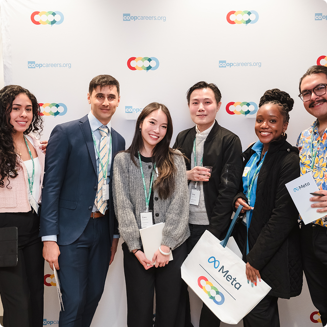 Group of six diverse young professionals smiling at a conference, standing in front of a backdrop with logos. Some hold bags and papers, one has a coffee cup.