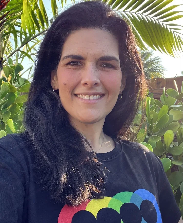 A woman with dark hair smiling outdoors amidst lush green tropical plants and palm leaves.