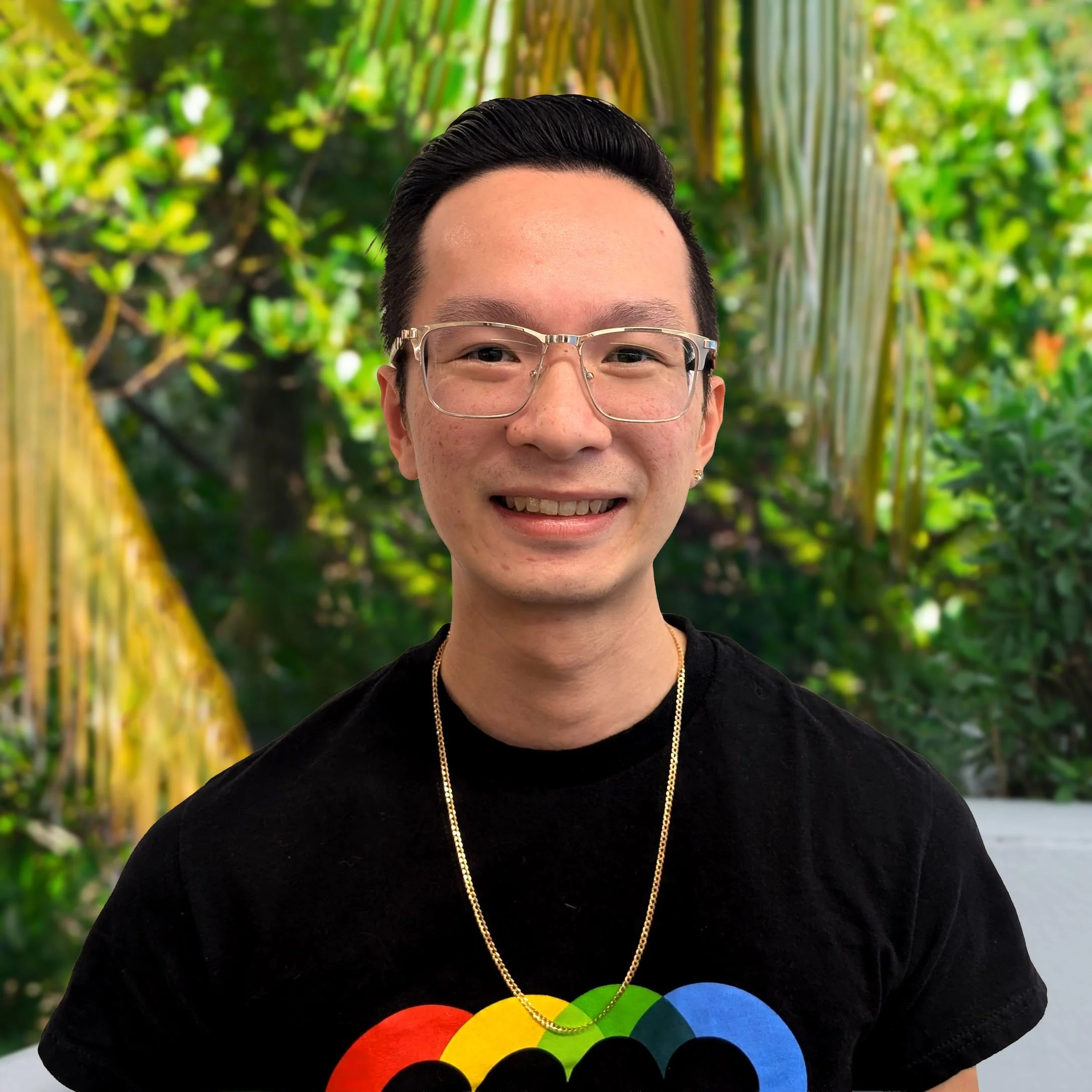 A young person with glasses smiling, wearing a black t-shirt with a rainbow-colored design, standing in outside in green nature setting