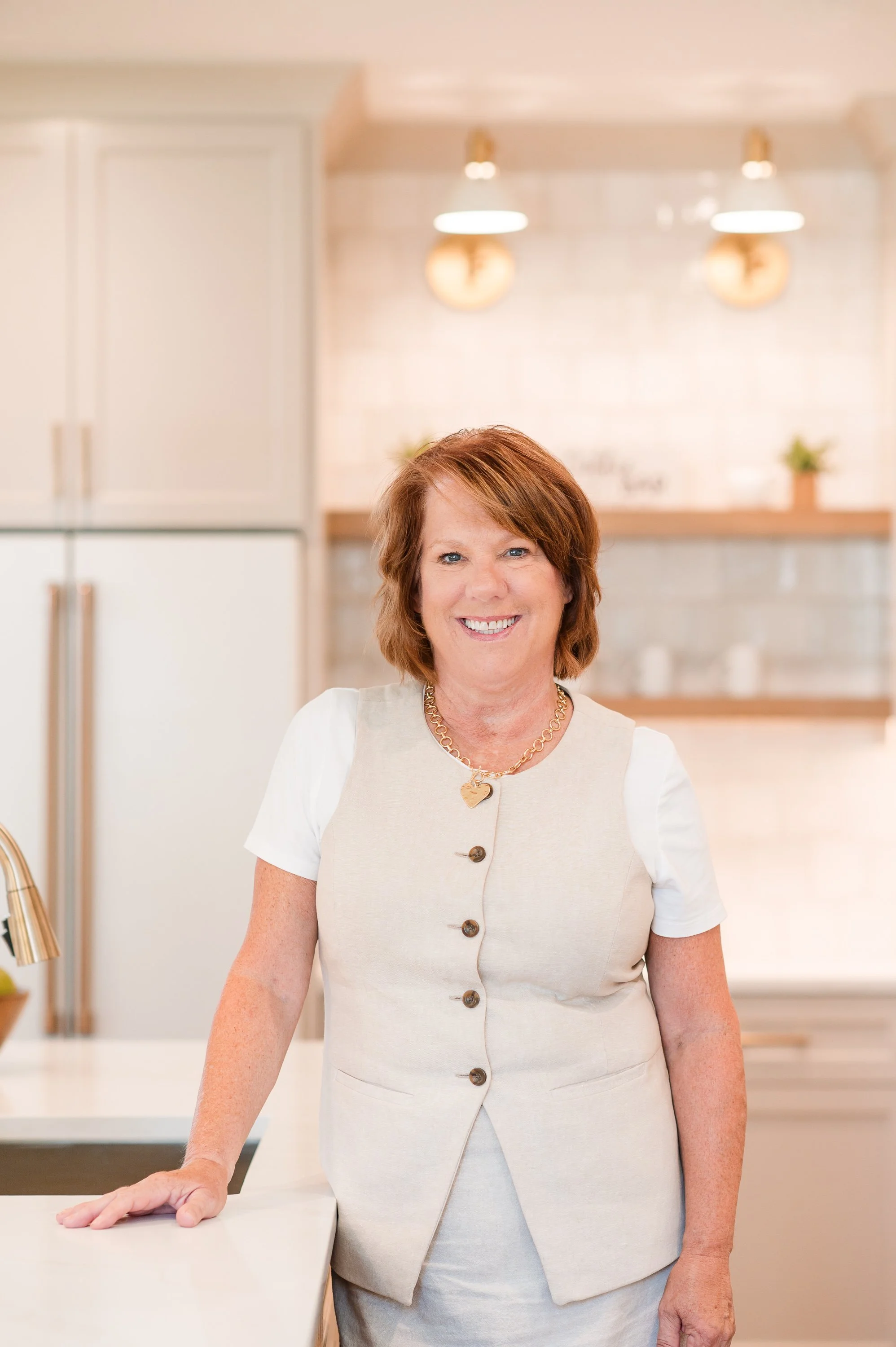 A smiling woman with short brown hair, wearing a light-colored vest and a white shirt, standing in a modern kitchen with beige and white tones. She has a gold necklace with a heart-shaped pendant and is resting her hand on a white kitchen countertop.