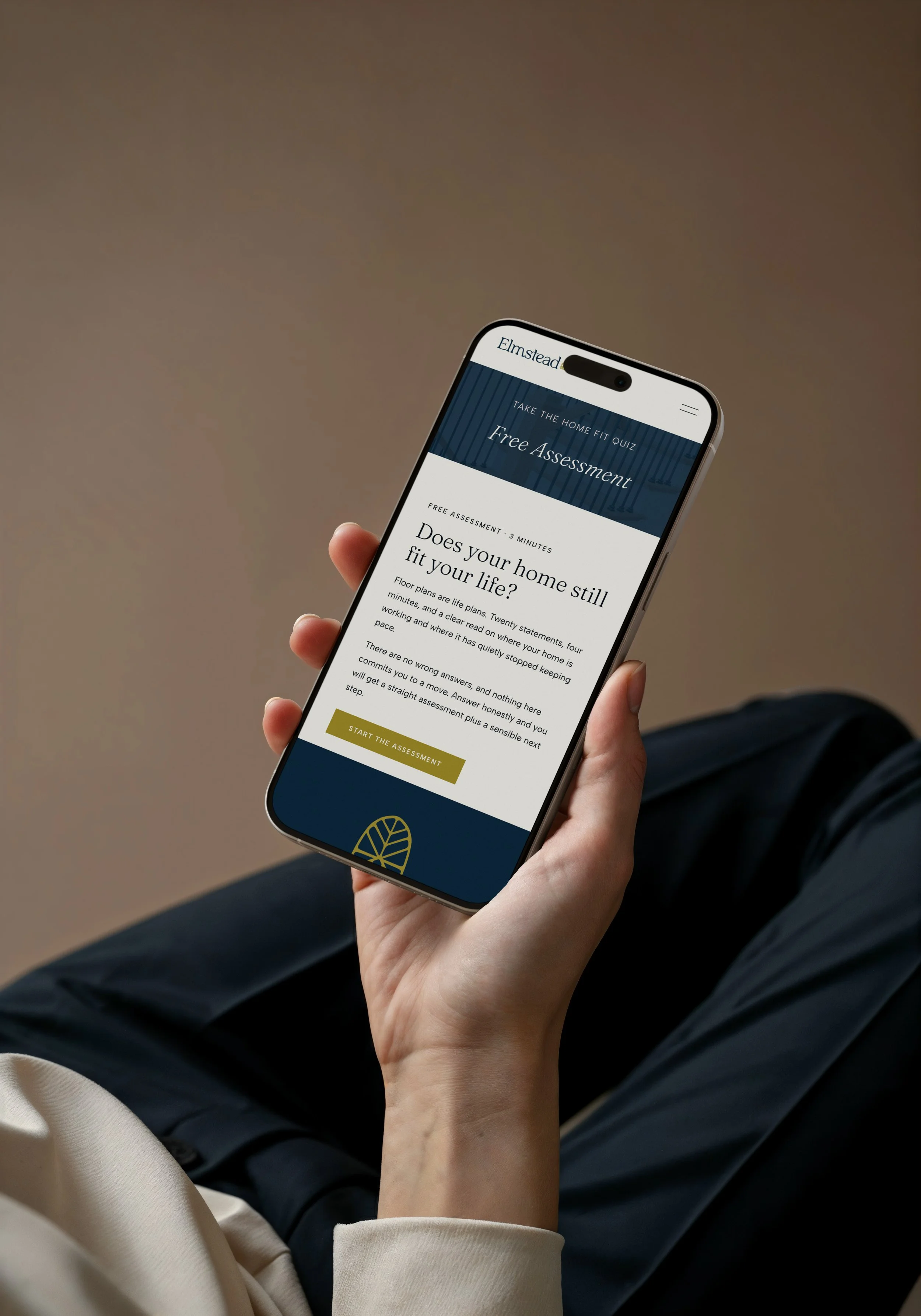 Person holding a smartphone with a website open on the screen, sitting with legs crossed, wearing dark pants and a cream-colored sleeve. The webpage is for a home fitness assessment.