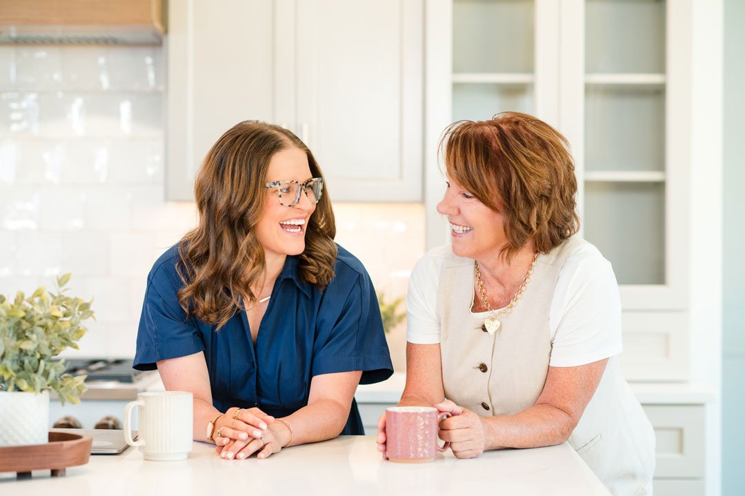 Two women, smiling and laughing, talking in a bright kitchen with white cabinets and a marble countertop.