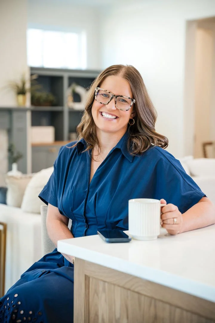 A woman with glasses, wearing a blue top, smiling and sitting at a kitchen counter with a coffee mug and smartphone.