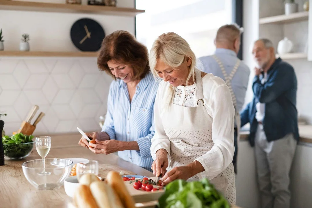 Four people in a kitchen, two women preparing food, two men talking in the background, with vegetables, wine glass, and kitchen decor visible.