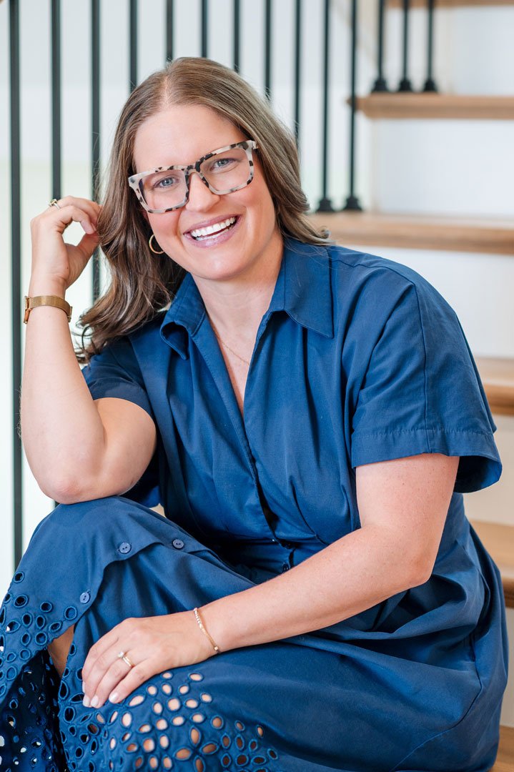A woman with shoulder-length light brown hair and glasses, smiling and sitting on stairs, wearing a blue dress with eyelet details.