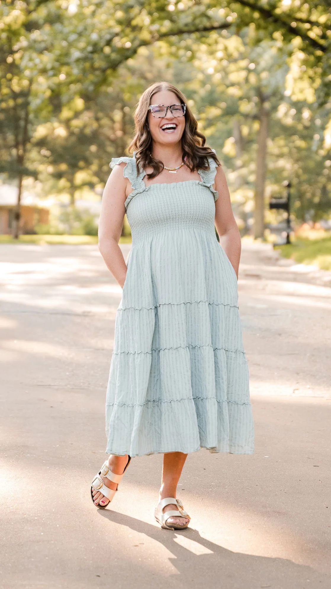 A woman with brown hair and glasses, smiling while walking outdoors on a sunny day. She is wearing a long, light blue, tiered dress, sandals, and jewelry, with trees in the background.