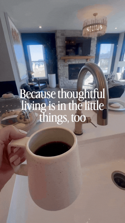 A person holding a mug of coffee in a modern kitchen with a living room in the background, featuring a stone fireplace, large windows, and contemporary furnishings, with an overlaid text saying, "Because thoughtful living is in the little things, too."