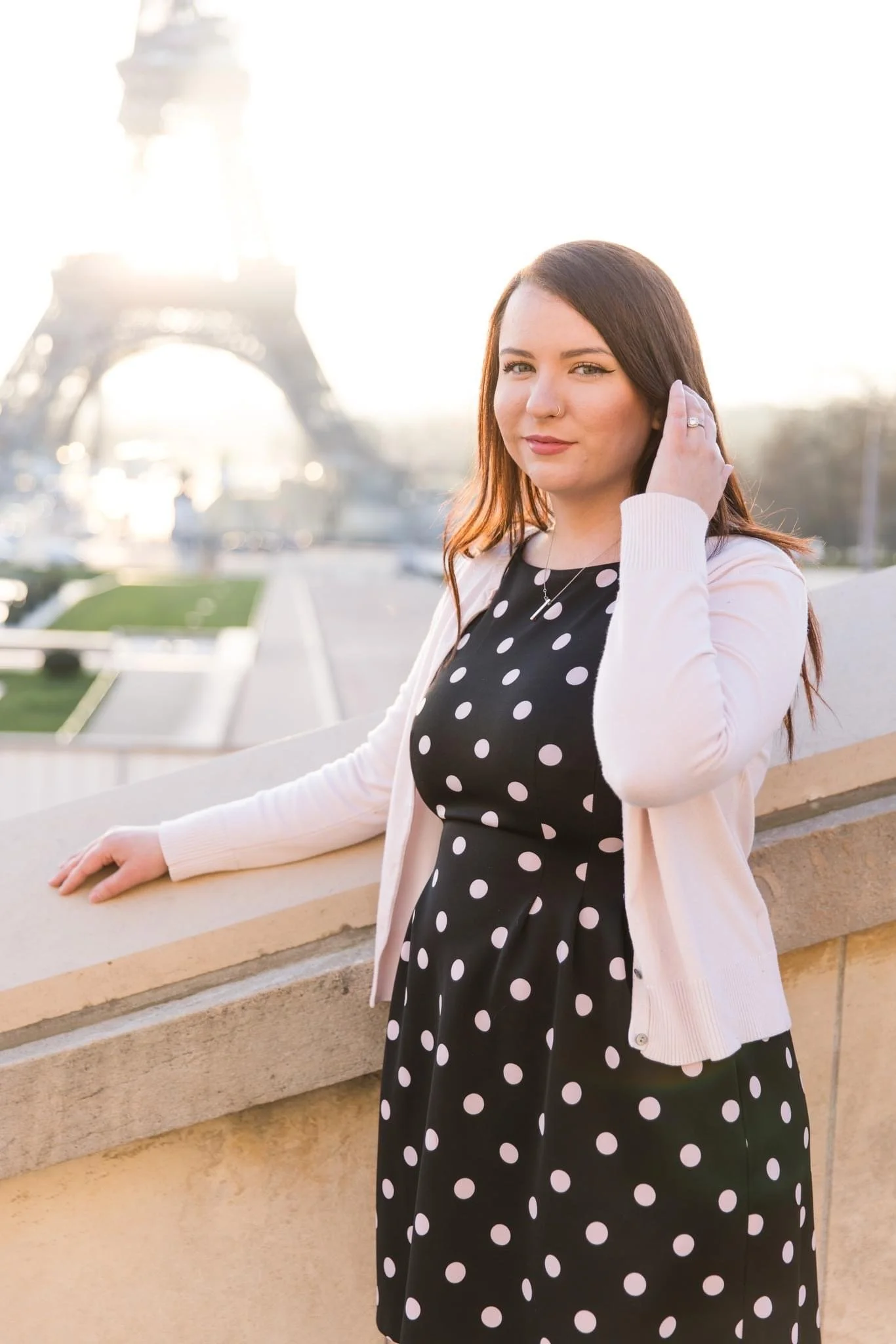 A woman with long brown hair wearing a black dress with white polka dots and a light pink cardigan, standing near a balcony with the Eiffel Tower in the background during sunset.