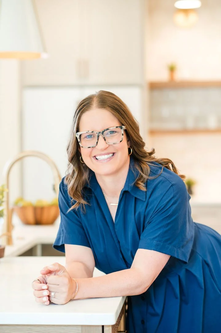 A smiling woman with brown hair, wearing glasses and a blue dress, leaning on a kitchen counter.