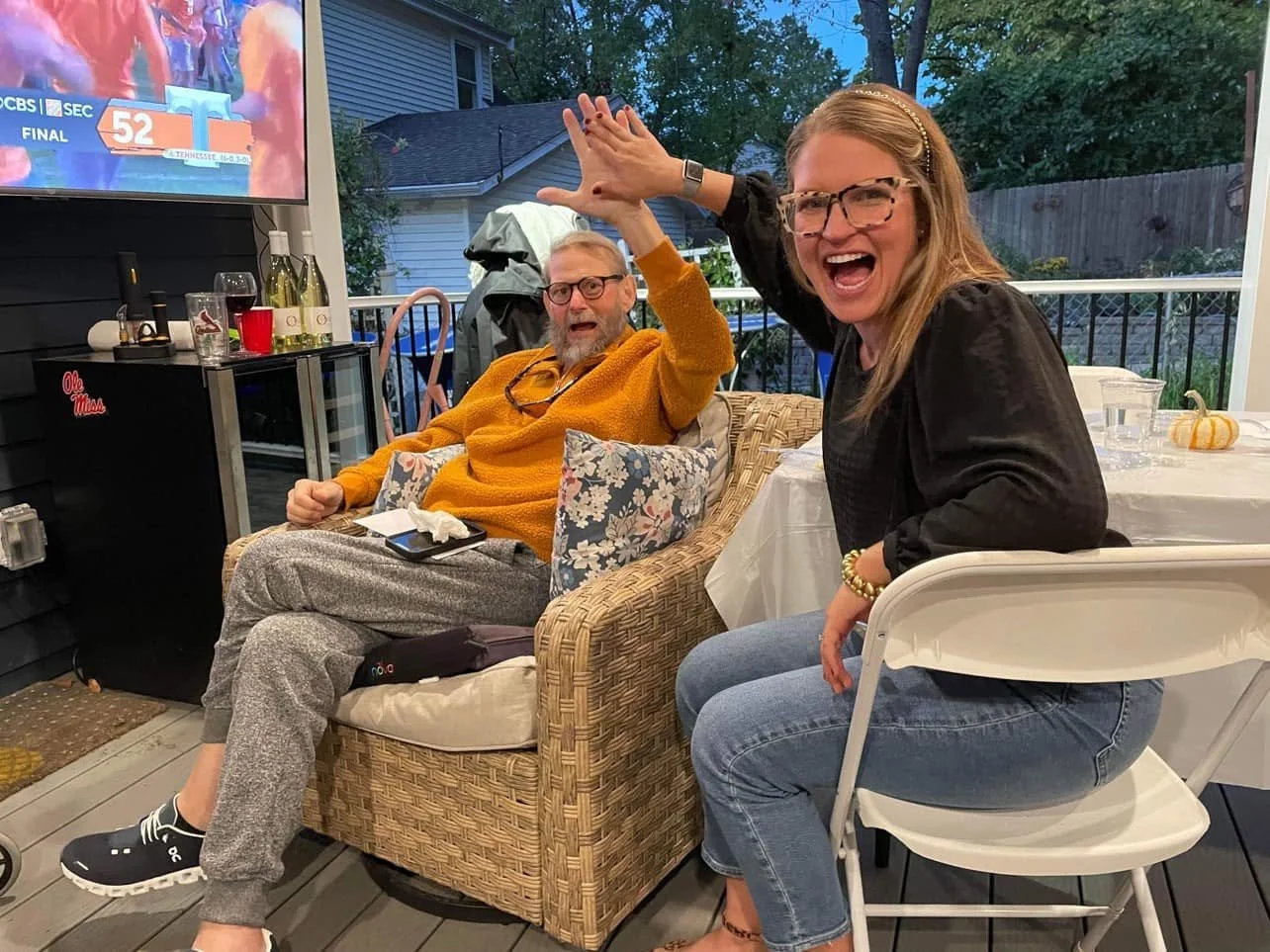 Two women outdoors on a patio during evening, one seated on a wicker chair wearing a mustard sweater and discussing with a woman sitting on a white folding chair wearing glasses, a black top, and blue jeans, both smiling and reaching towards each other.