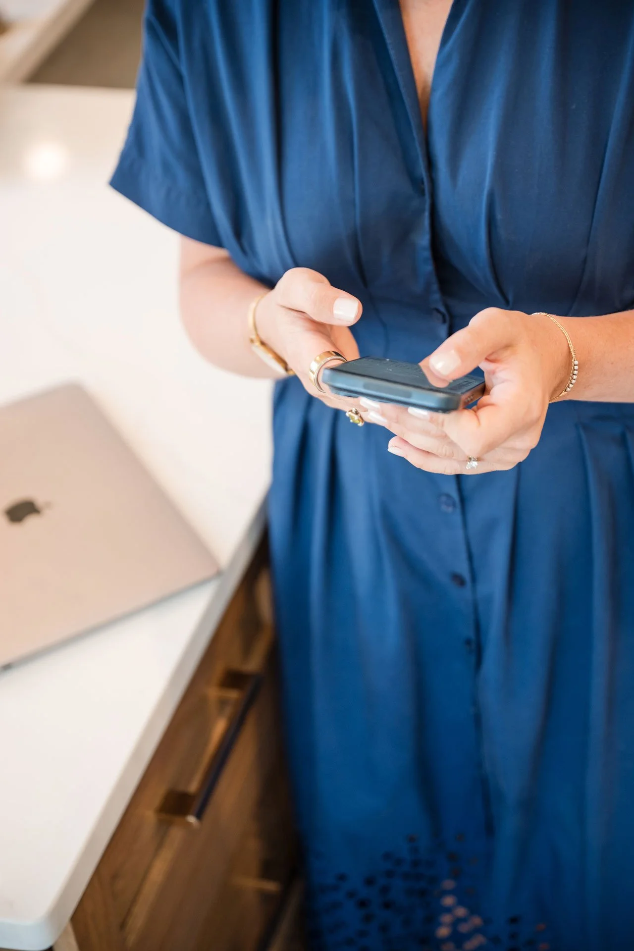 Person wearing a blue dress using a smartphone, with a closed laptop on a white table nearby.
