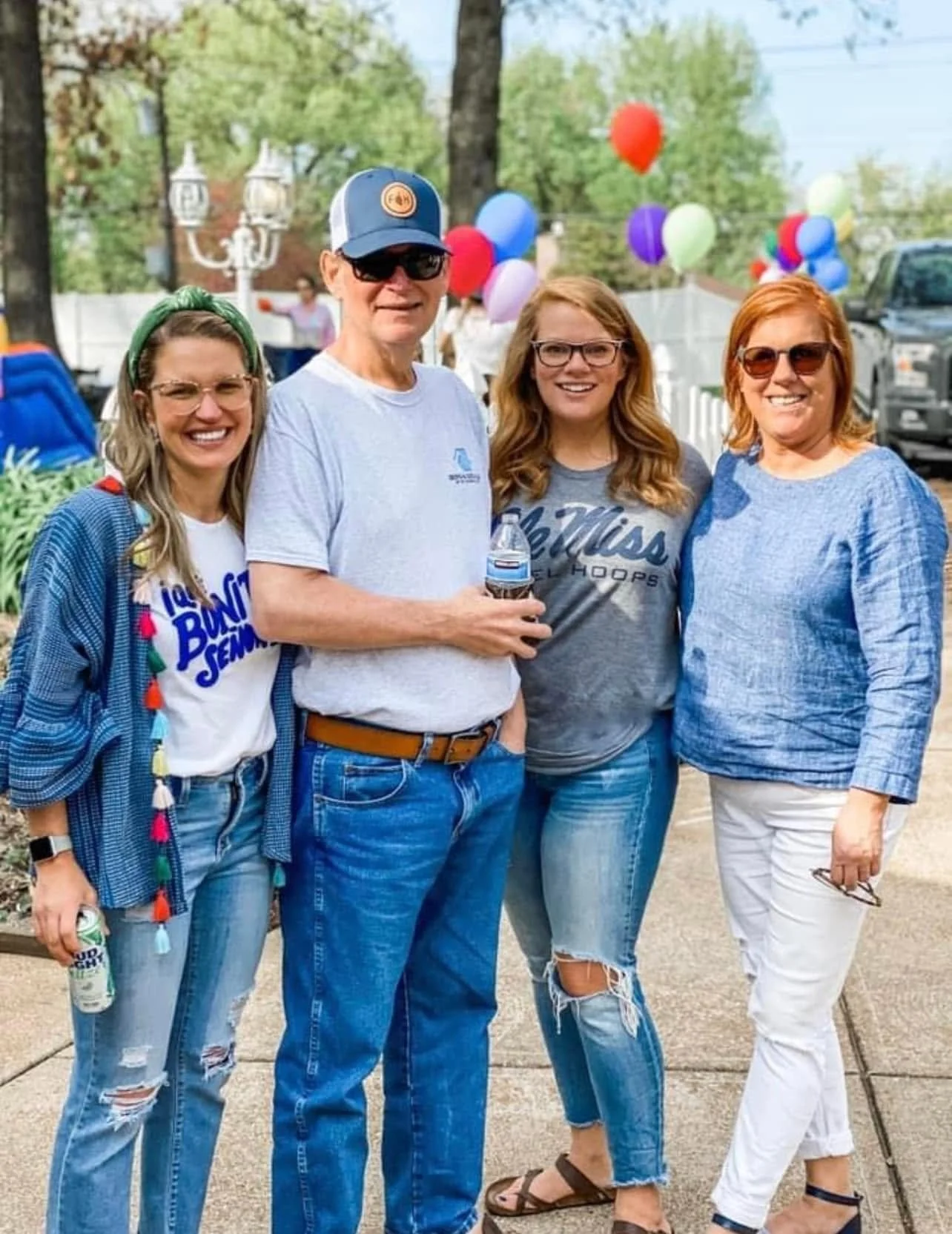 Four women and one man smiling outdoors at a party with balloons in the background. The women are wearing casual clothing with jeans or white pants, and the man is holding a water bottle and wearing sunglasses and a cap.