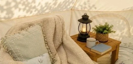 Cozy living space with a beige plush sofa, a throw pillow, and a small wooden side table holding a lantern, a potted plant, a mug, and a book.