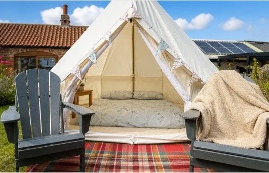 Canvas tent set up outdoors with two dark gray chairs, a blanket on one chair, and a rug on the grass, against a background of a brick house and solar panels.